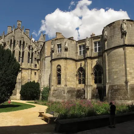 Un jardin public, un chemin de gravier et, derrière les arbres, la façade et la tour du palais.